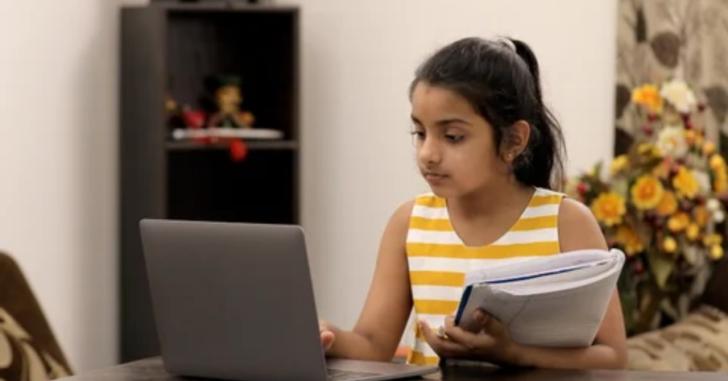 Indian schoolgirl doing homework on laptop while holding a notebook